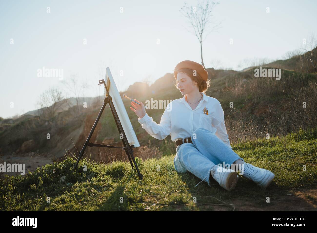 Full body of young Woman in beret and stylish clothes sitting on grassy lawn near hills while painting picture with brush on canvas on easel in sunny Stock Photo