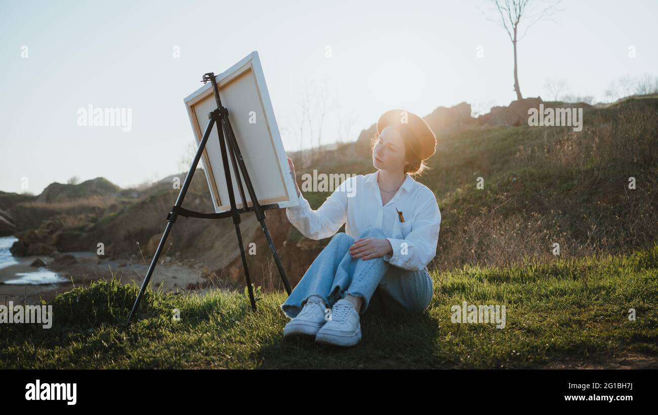 Full body of young Woman in beret and stylish clothes sitting on grassy lawn near hills while painting picture with brush on canvas on easel in sunny Stock Photo