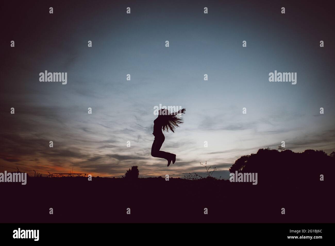 Low angle side view full body silhouette of unrecognizable female with outstretched arms and flying hair leaping above ground at sundown Stock Photo