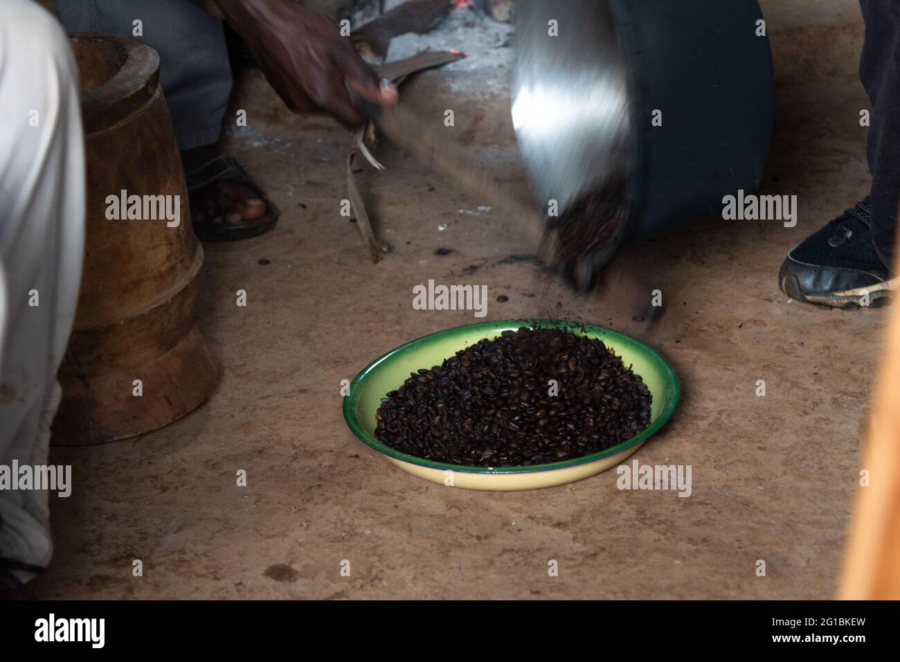 The roasted coffee beans are poured out of the pot into a ceramic bowl. A hand hits the pot with a stick Stock Photo