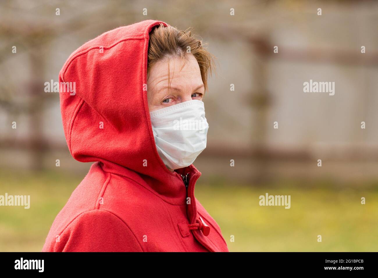 An elderly woman with brown hair and blue eyes in a red coat and hood in a protective safe medical mask, close-up portrait. Stock Photo