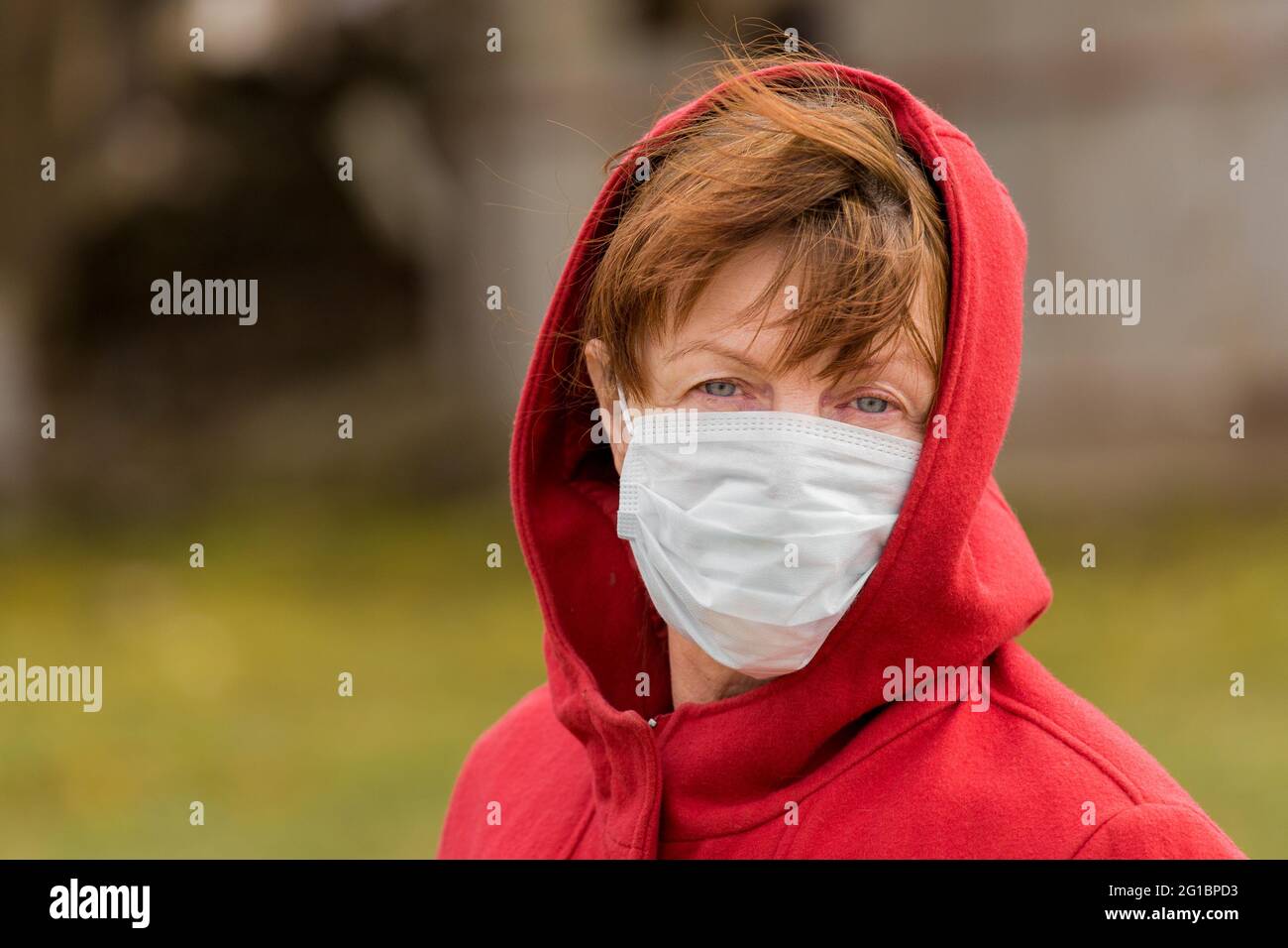 An elderly woman with brown hair and blue eyes in a red coat and hood in a protective safe medical mask, close-up portrait. Stock Photo