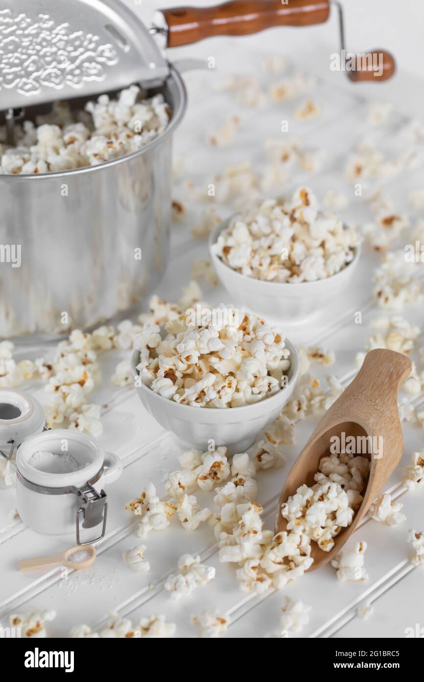 Popcorn pan maker, white bowls and wooden scoop full of popcorn. Messy food scene, in white. Retro, vintage ambience. Stock Photo