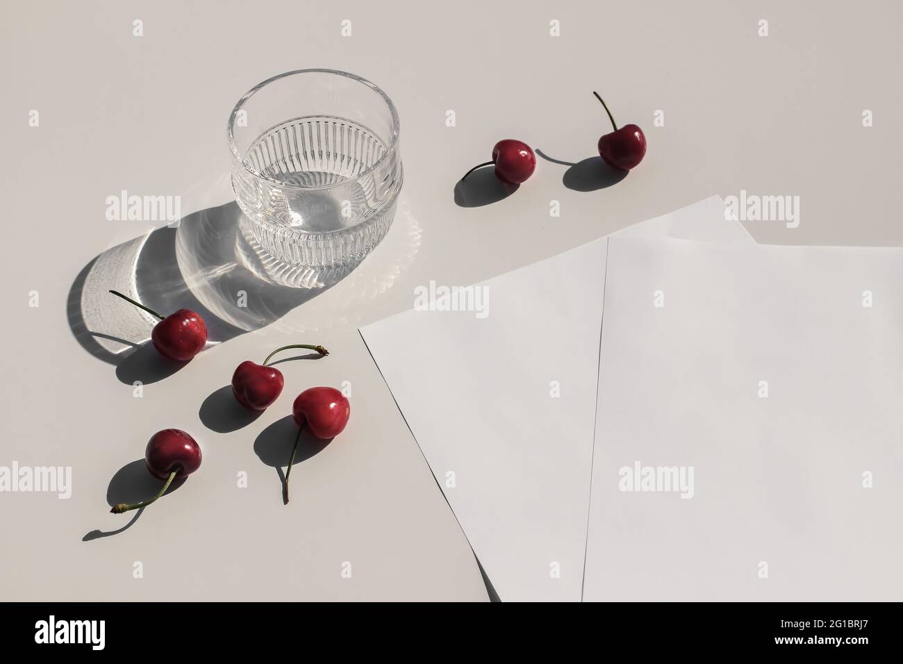 Summer food still life composition. Fresh red cherries fruit on beige table background. Stationery mock up scene. Blank paper pages. Glass of water Stock Photo