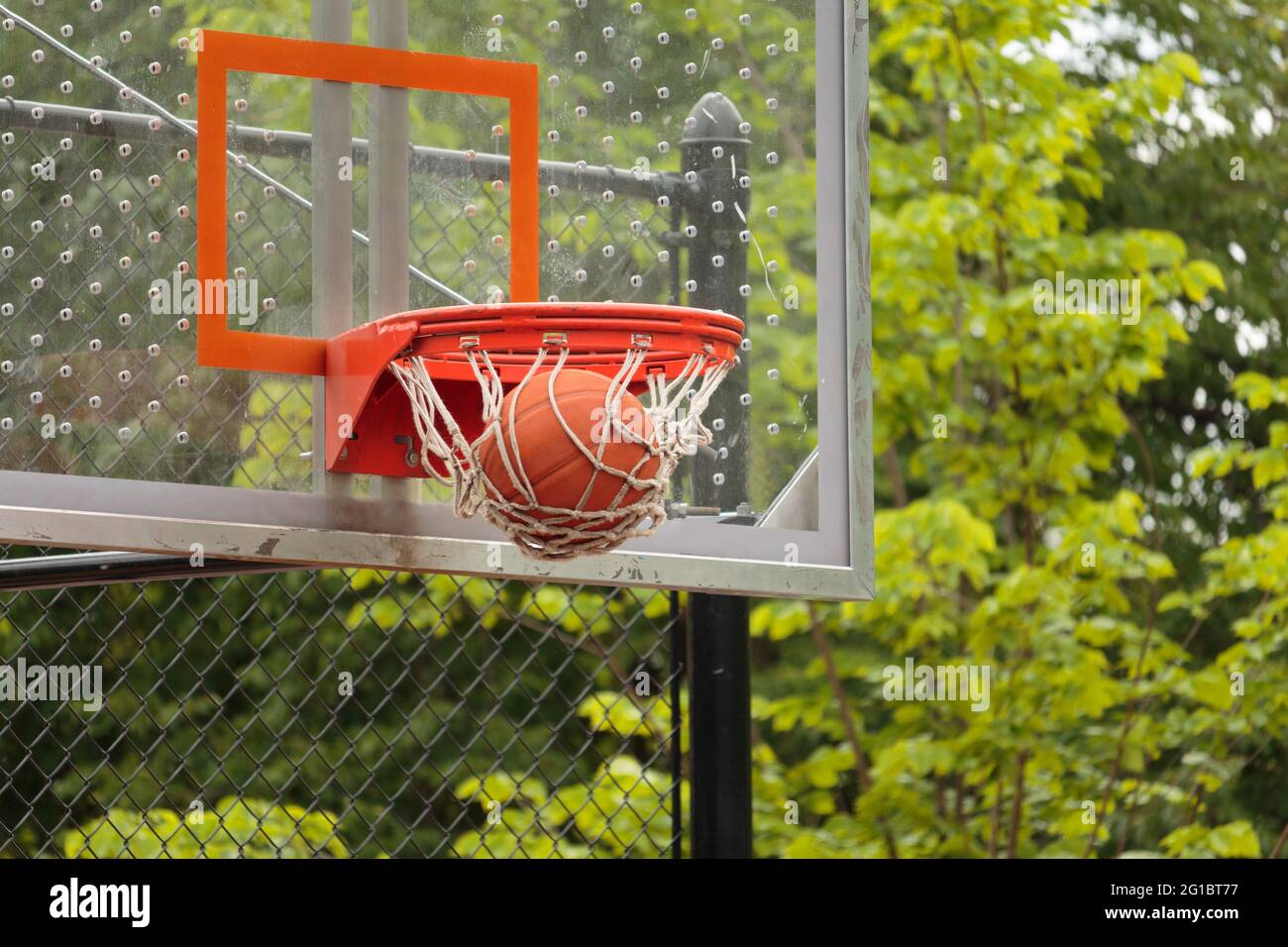 close up of a basketball going through the net of the basketball hoop with glass backboard visible and trees in background, a score in basketball game Stock Photo
