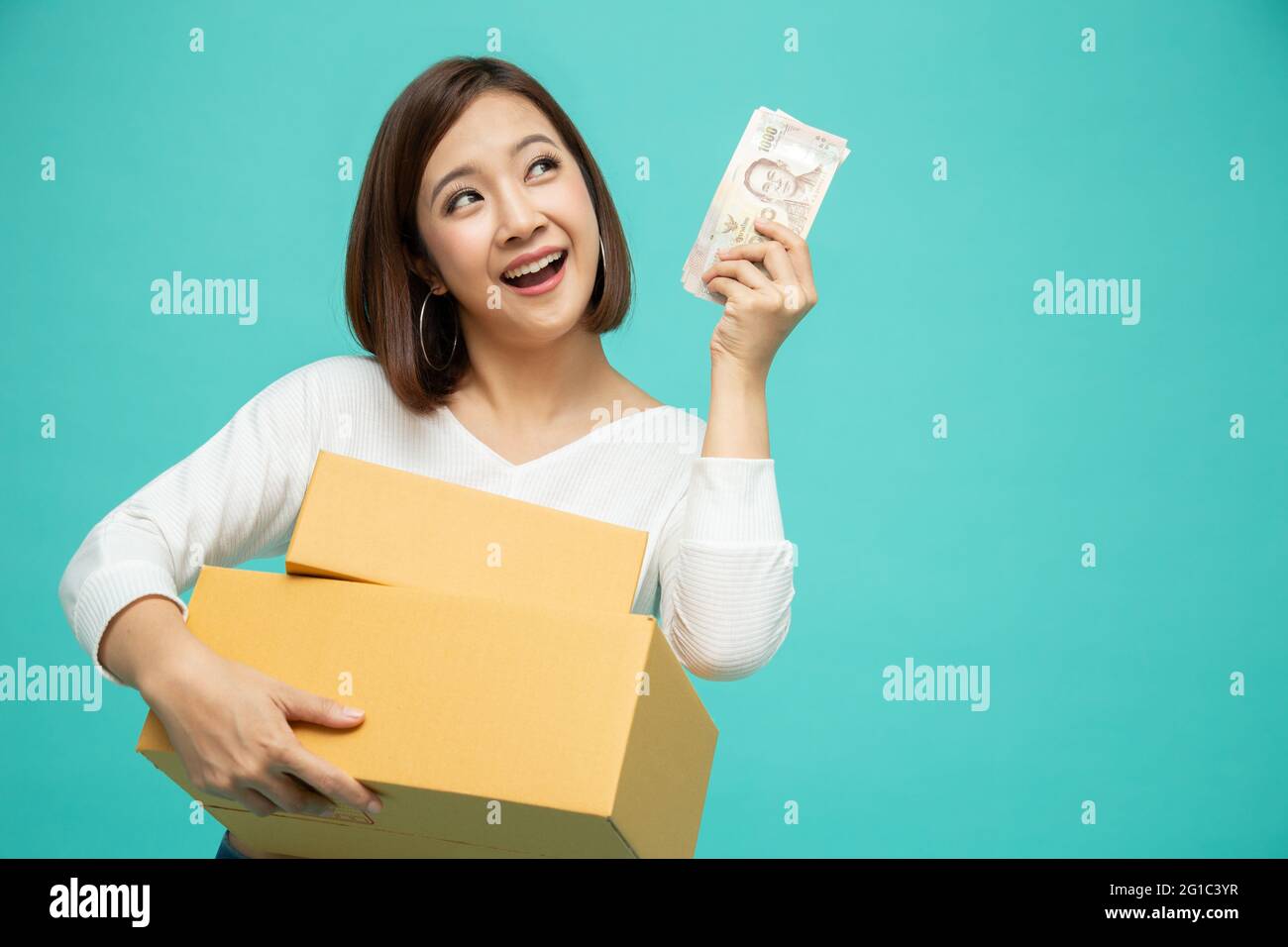 Happy Asian woman holding package parcel box and celebrating with money banknotes thai baht isolated on green background, Delivery courier and shippin Stock Photo