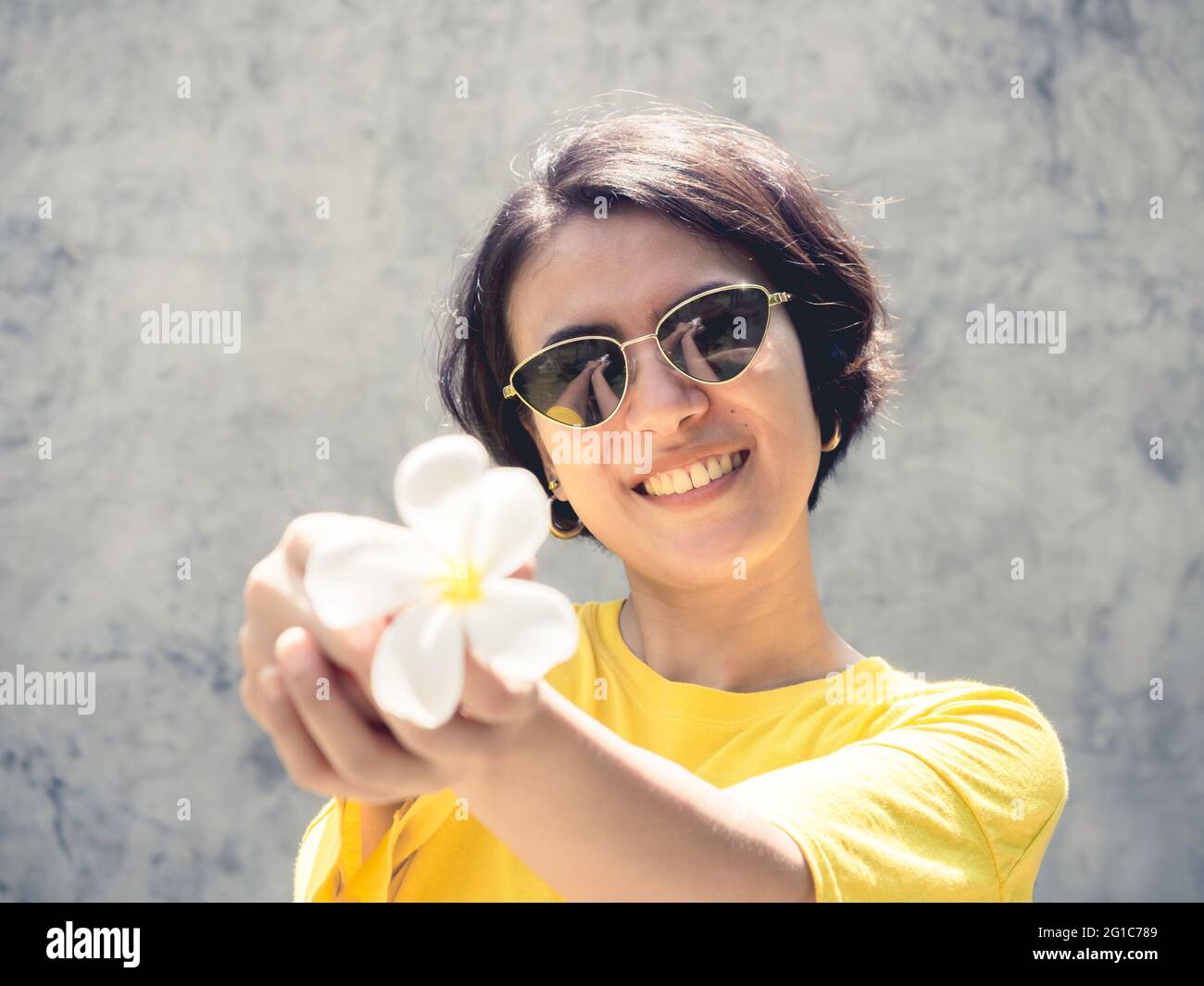 Summertime. Smile with the sunshine. Beautiful Asian woman short hair wearing sunglasses and yellow shirt showing the white plumeria flower on concret Stock Photo