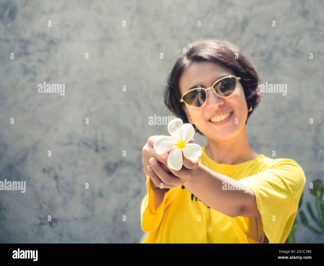 Summertime. Smile with the sunshine. The white plumeria flower on hand of beautiful Asian woman short hair wearing sunglasses and yellow shirt on conc Stock Photo