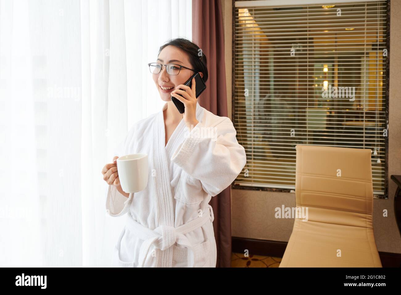 Happy young woman in bathrobe drinking morning coffee and talking on phone Stock Photo