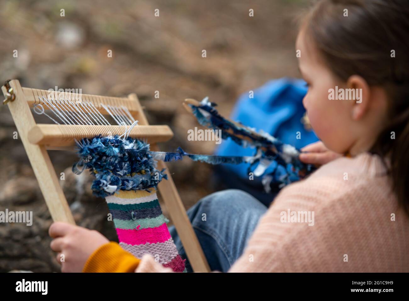 Small girl with hand loom sitting outdoors in city park, learning group education concept. Stock Photo