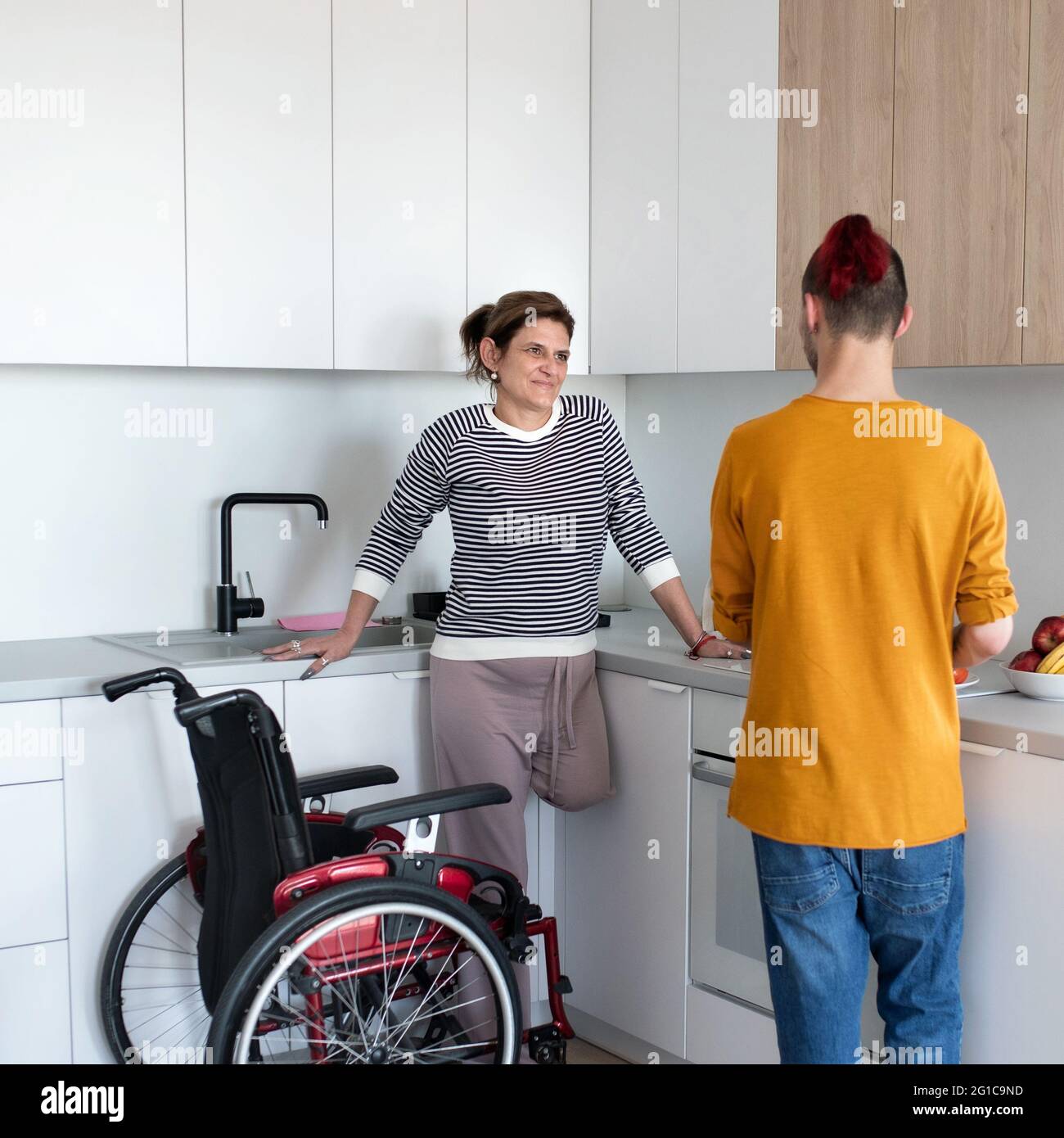 Disabled mature woman in wheelchair talking to son in kitchen indoors at home. Stock Photo