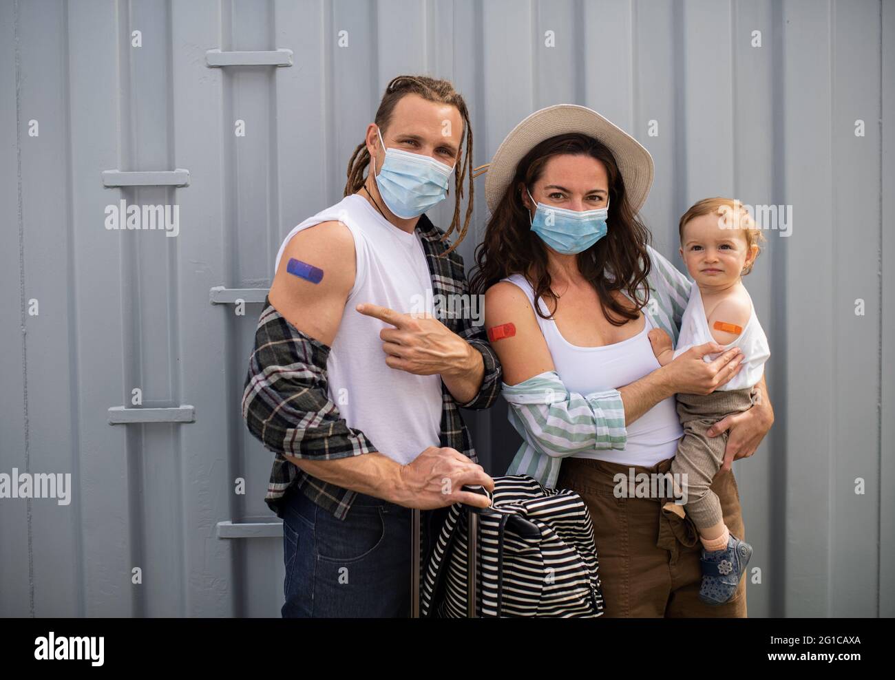 Portrait of young family with small baby after covid-9 vaccination, pointing to plaster on arm and looking at camera. Stock Photo