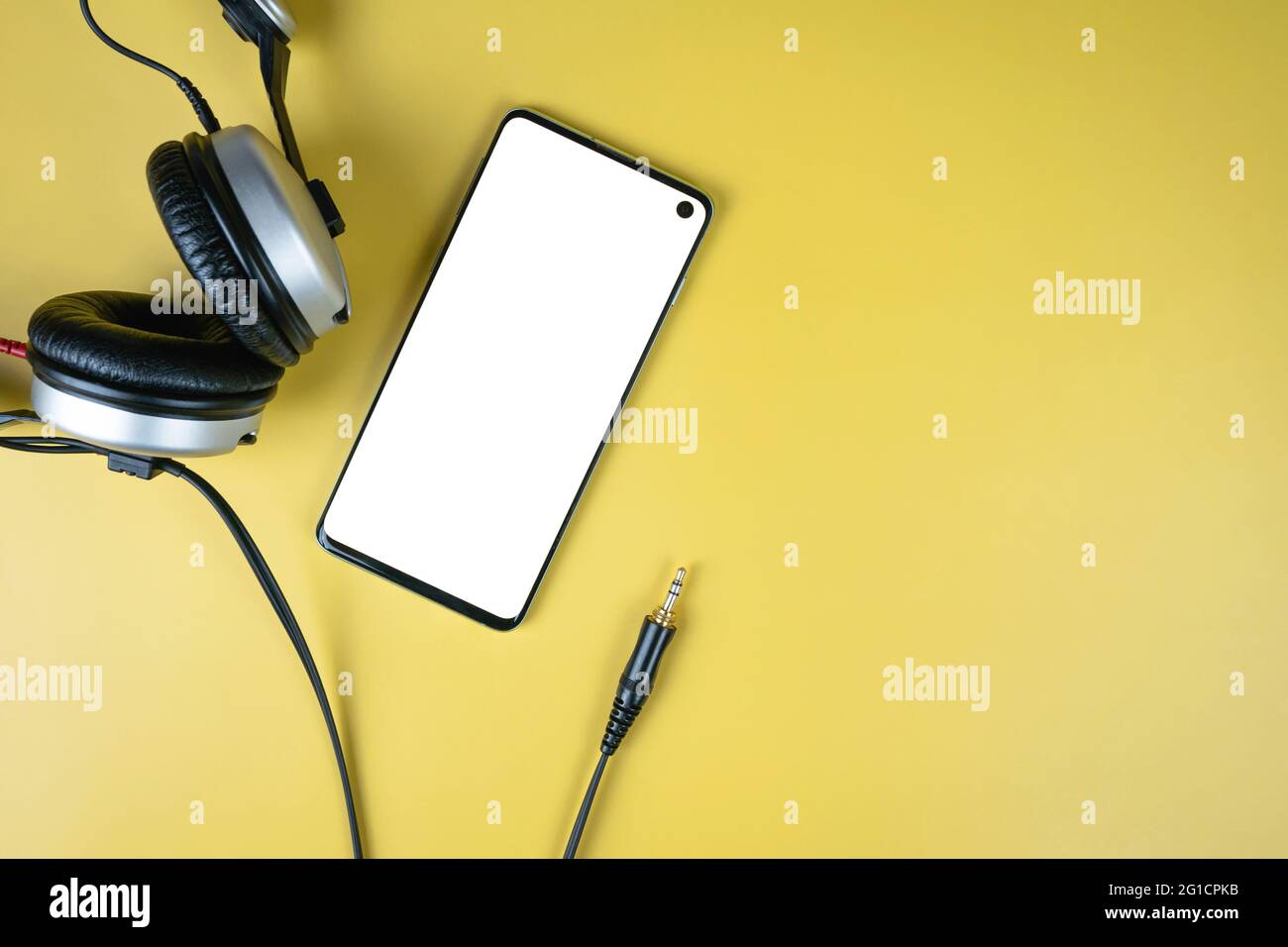 Modern smartphone with white mockup on screen with closed headphones with jack on yellow background with copy space, top view. Template for music apps Stock Photo