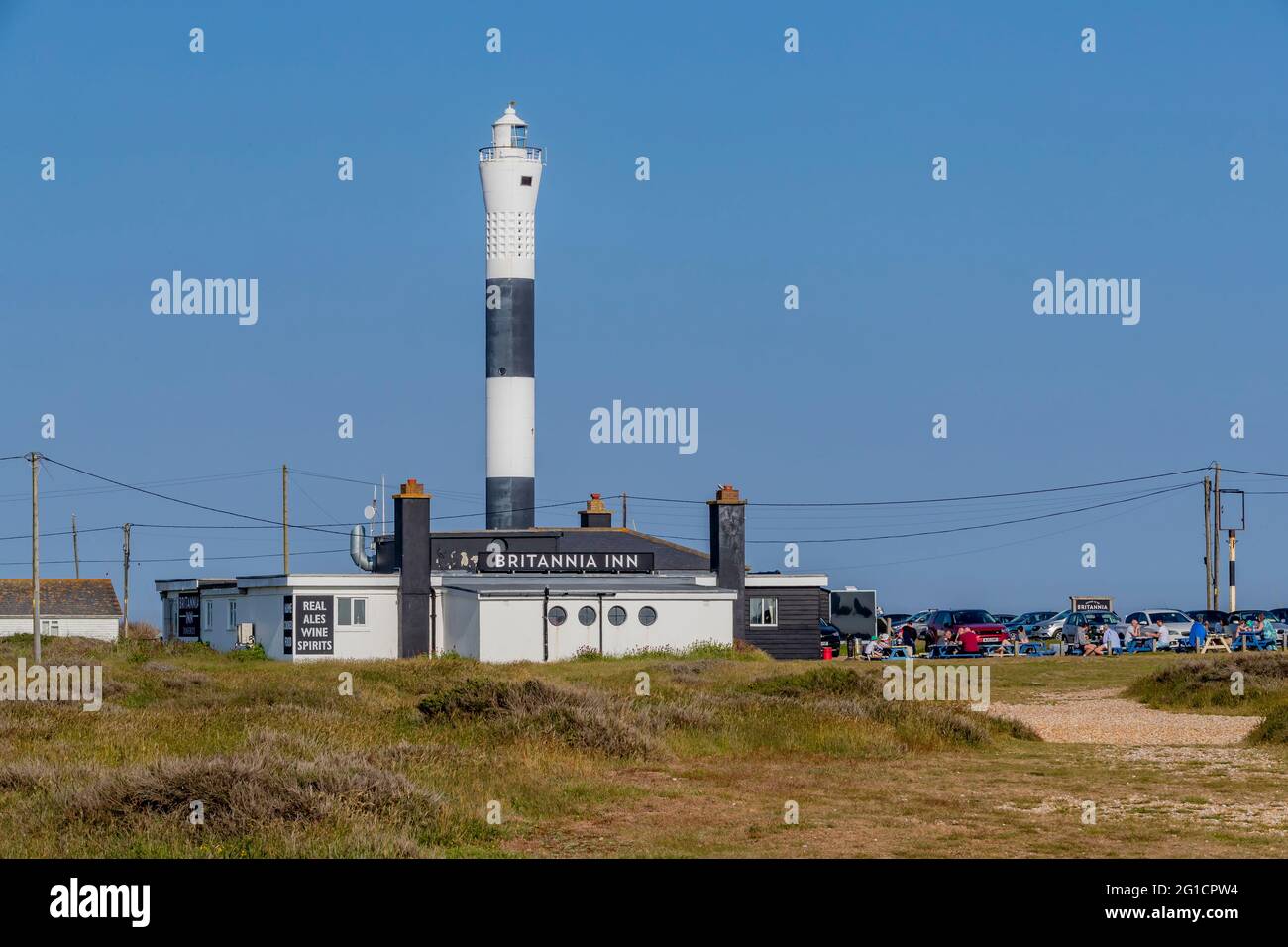 The Britannia Inn with prople sat outside enjoying the sunshine and a drink with the New Lighthouse in the background at Dungeness, Kent, England, UK. Stock Photo