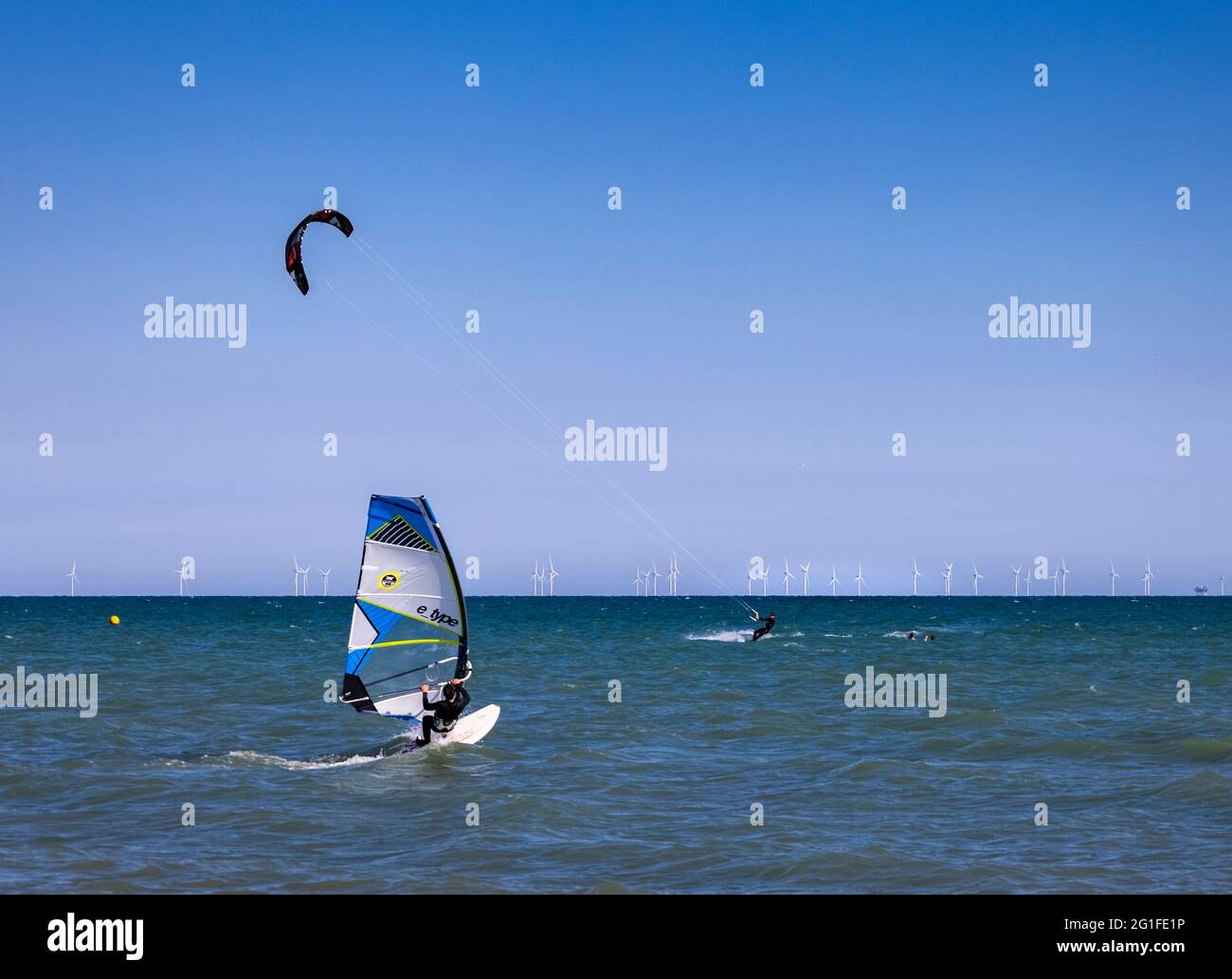 With a strong wind blowing two guys take to the ocean. One on a kiteboard and the other windsurfing. In the background it turns the wind farm turbines Stock Photo