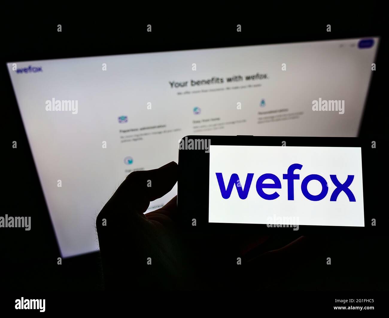 Person holding mobile phone with logo of digital insurance company wefox Holding AG on screen in front of business web page. Focus on phone display. Stock Photo