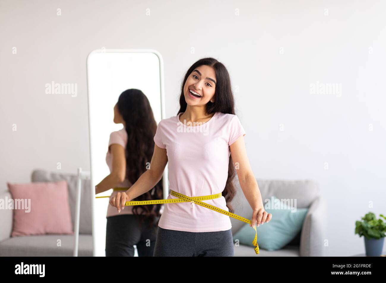 Indian woman measuring her waist with tape in front of mirror, showing results of slimming diet or liposuction at home Stock Photo