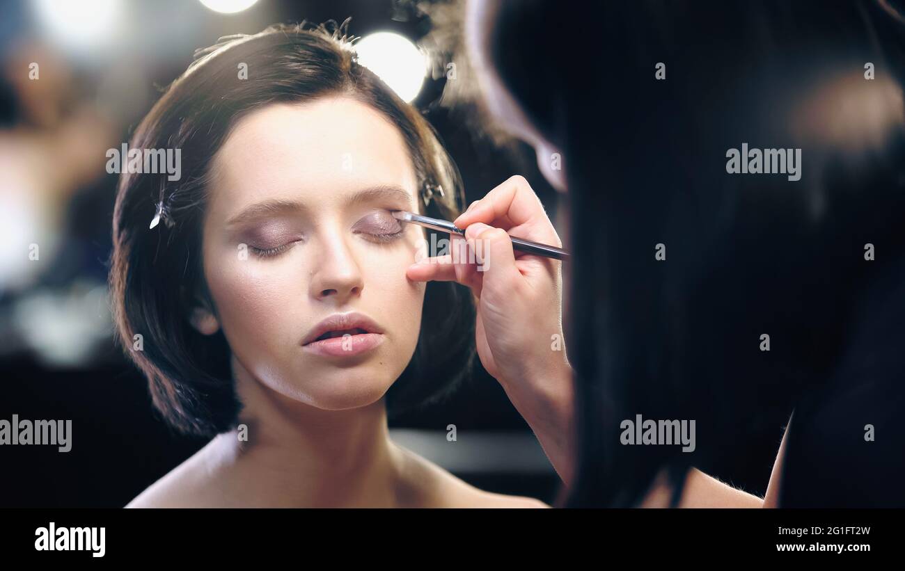 makeup artist applying eye shadow with cosmetic brush on young pretty model with closed eyes Stock Photo