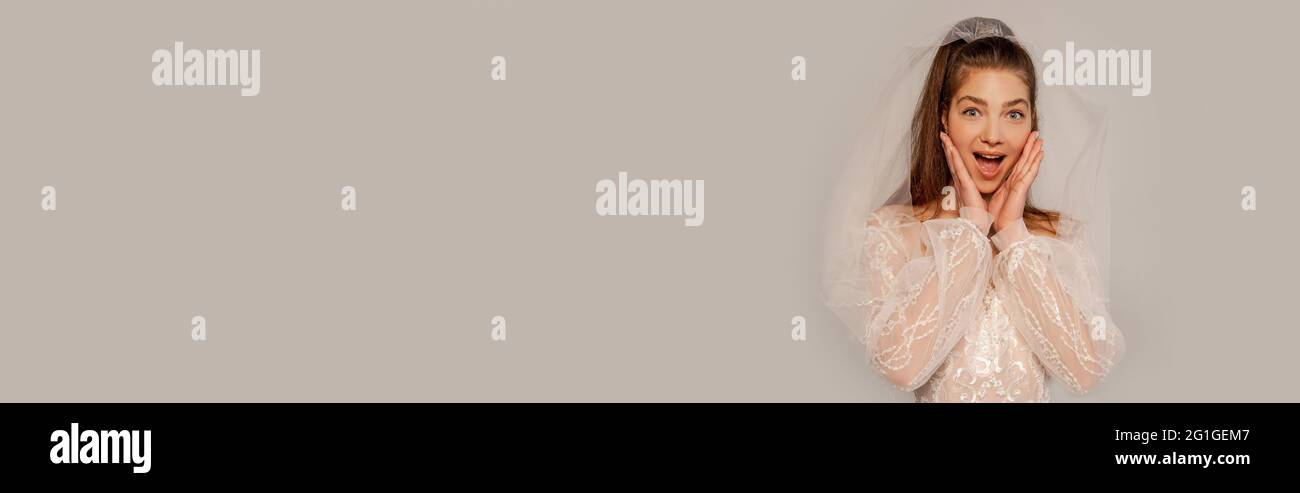astonished woman in wedding dress touching face isolated on grey, banner Stock Photo