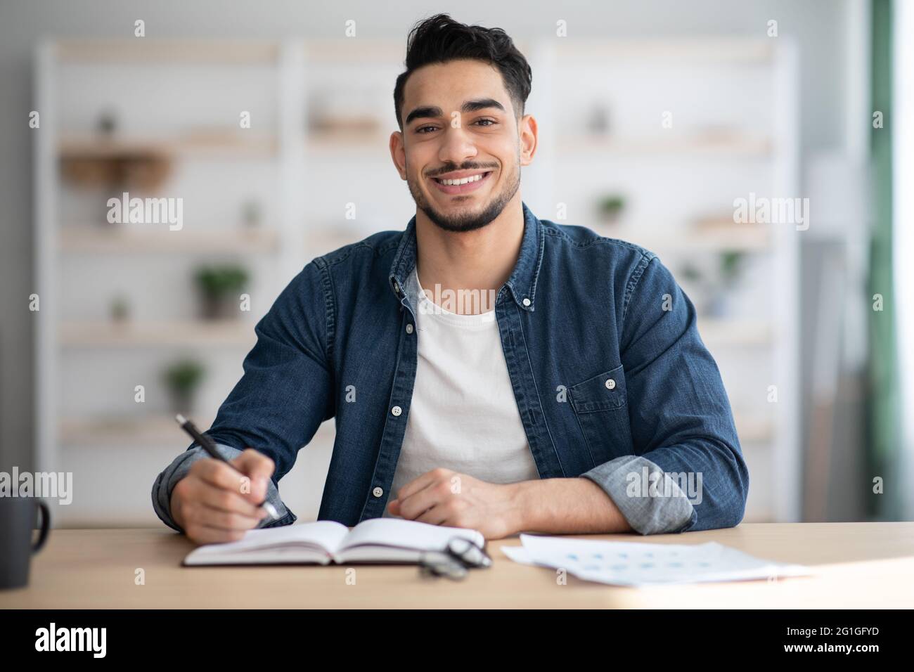 Cheerful arab guy in casual sitting at workdesk, taking notes Stock Photo