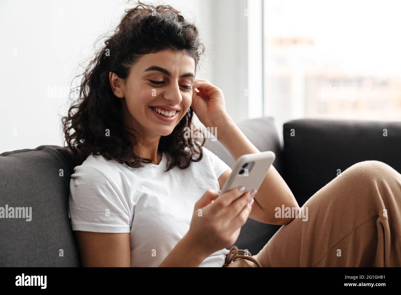 Happy brunette young woman using mobile phone while sitting on sofa at ...