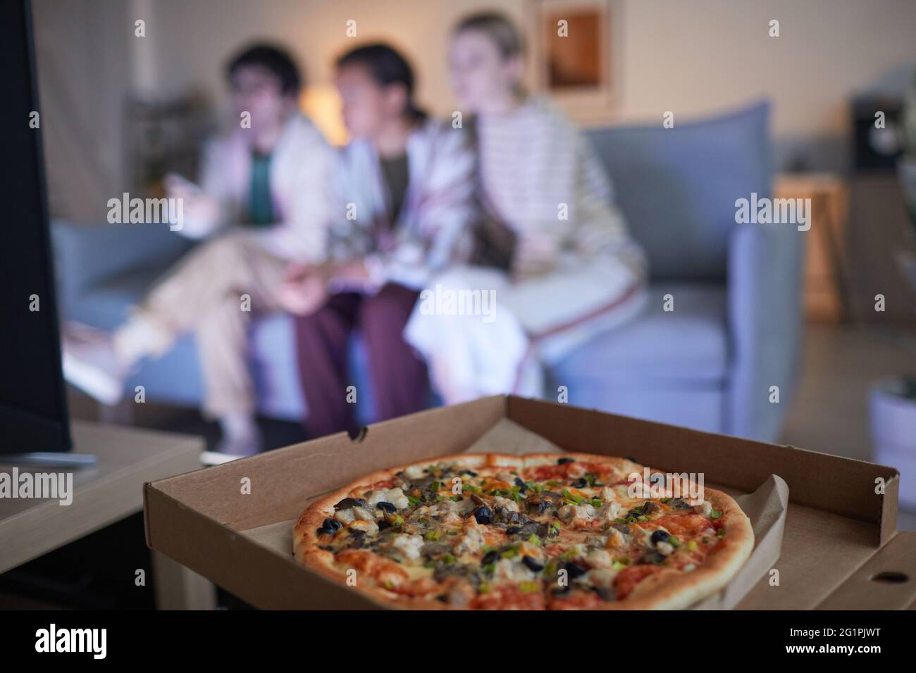Blurred group of friends watching TV at home lit by blue light, focus on pizza in foreground, copy space Stock Photo