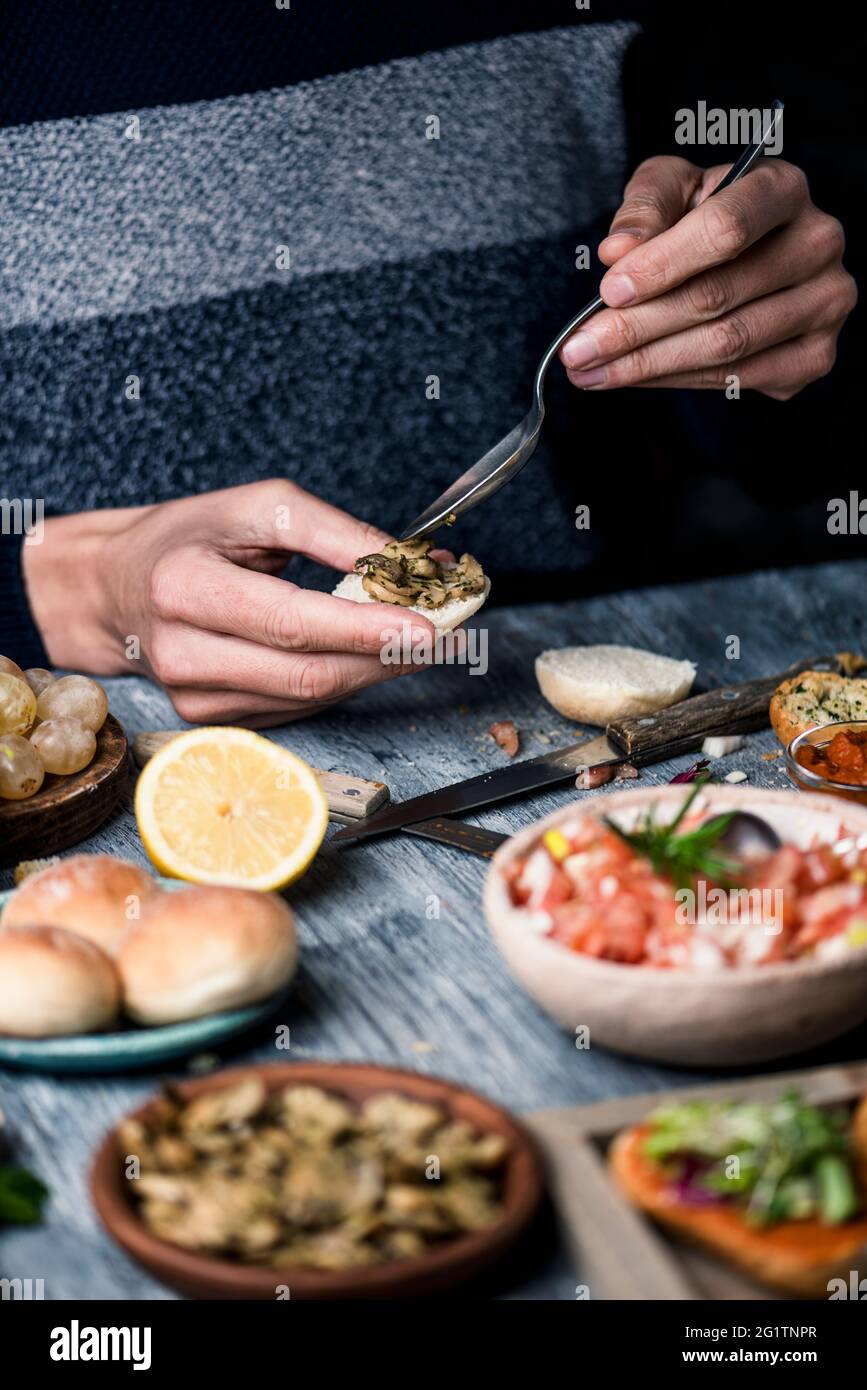 a young caucasian man, sitting at a gray rustic wooden table, puts some mushrooms fried with garlic and parsley on a halved bread bun Stock Photo