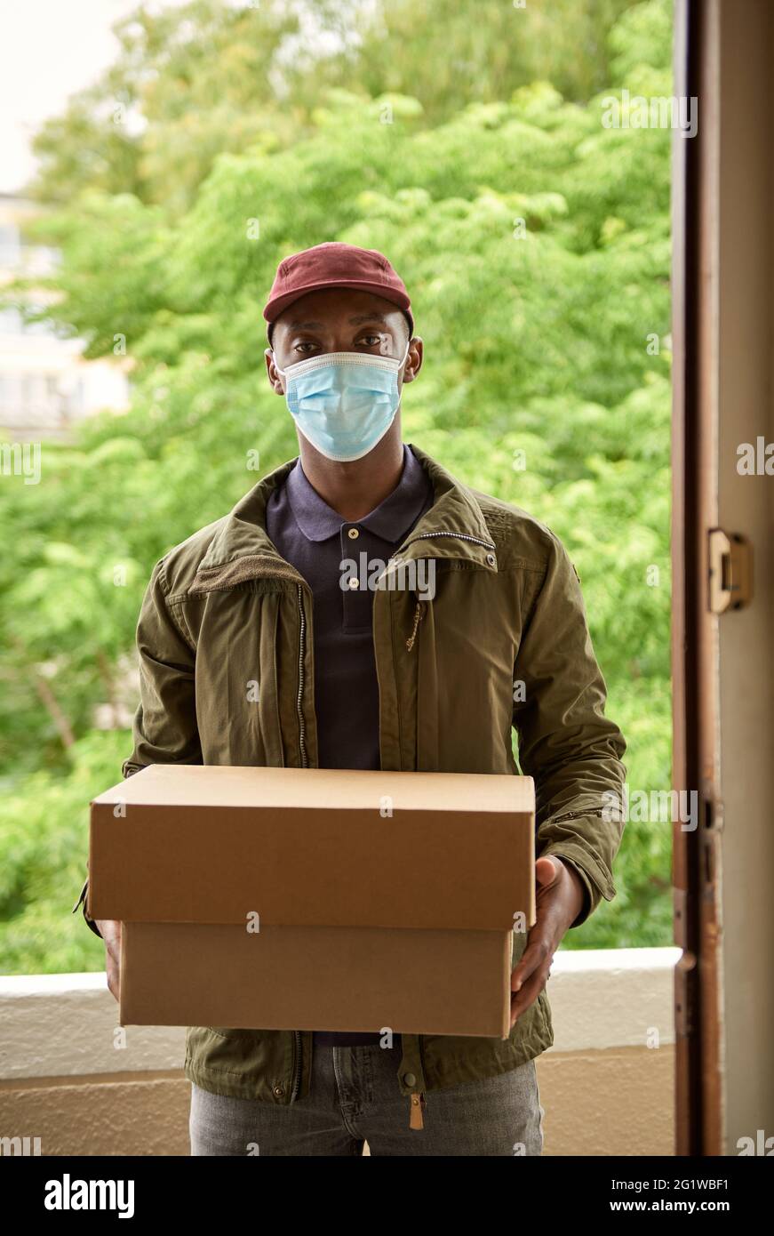 African courier in a mask standing with packages at a door Stock Photo