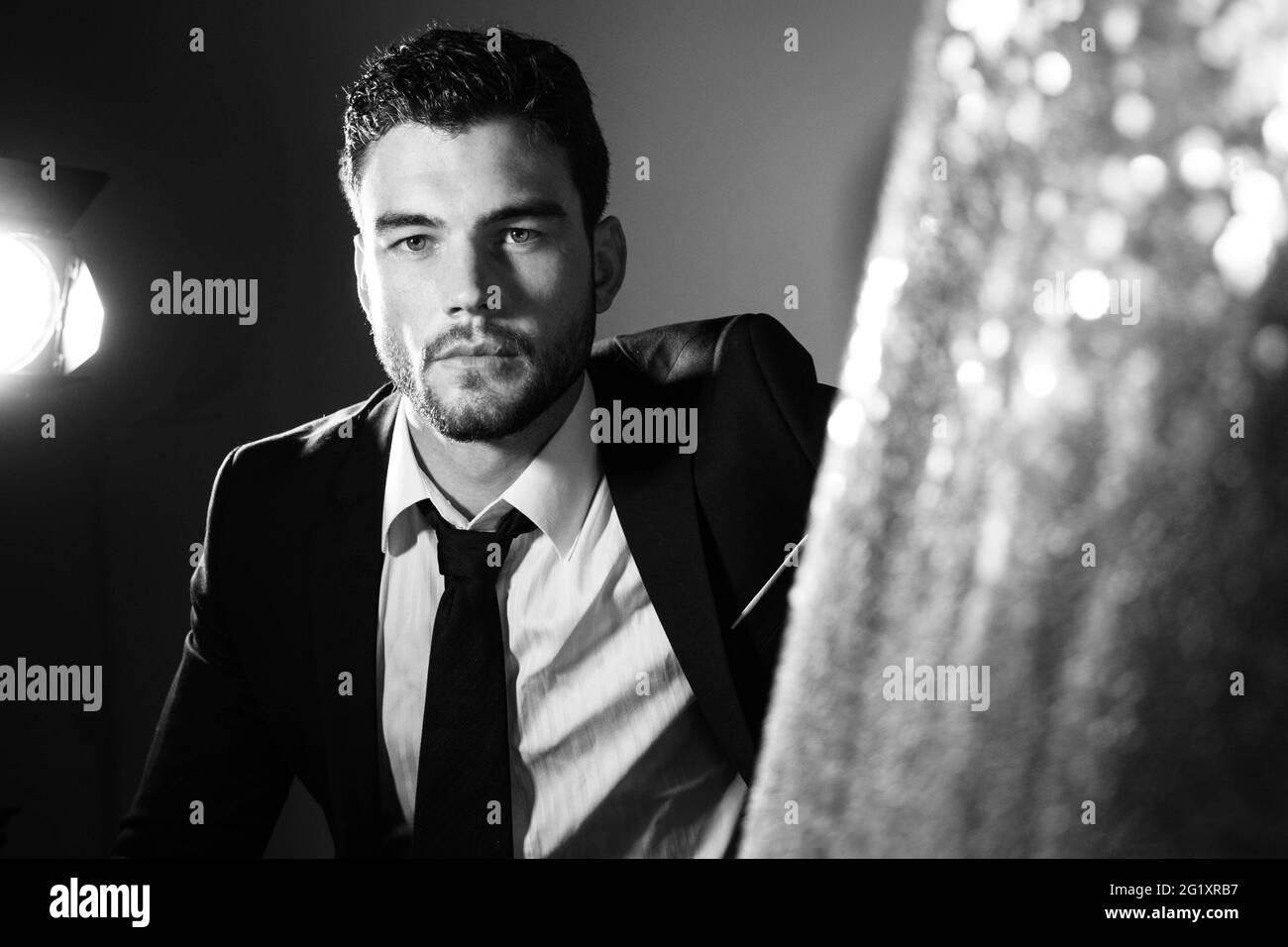 Attractive man wearing suit sitting next to glitter curtain, looking at camera with spotlight in background Stock Photo