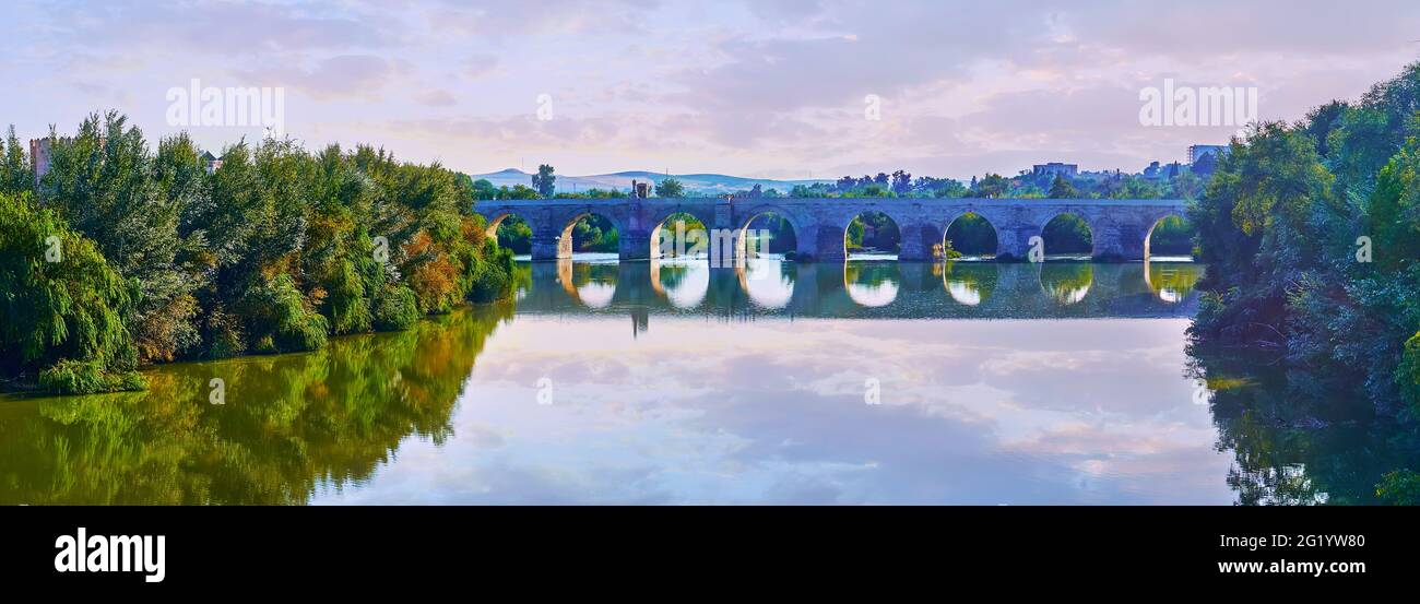Panorama of the antique pedestrian Puente Romano (Roman Bridge) across Guadalquivir River with reflection and lush thickets on the river's banks, Cord Stock Photo