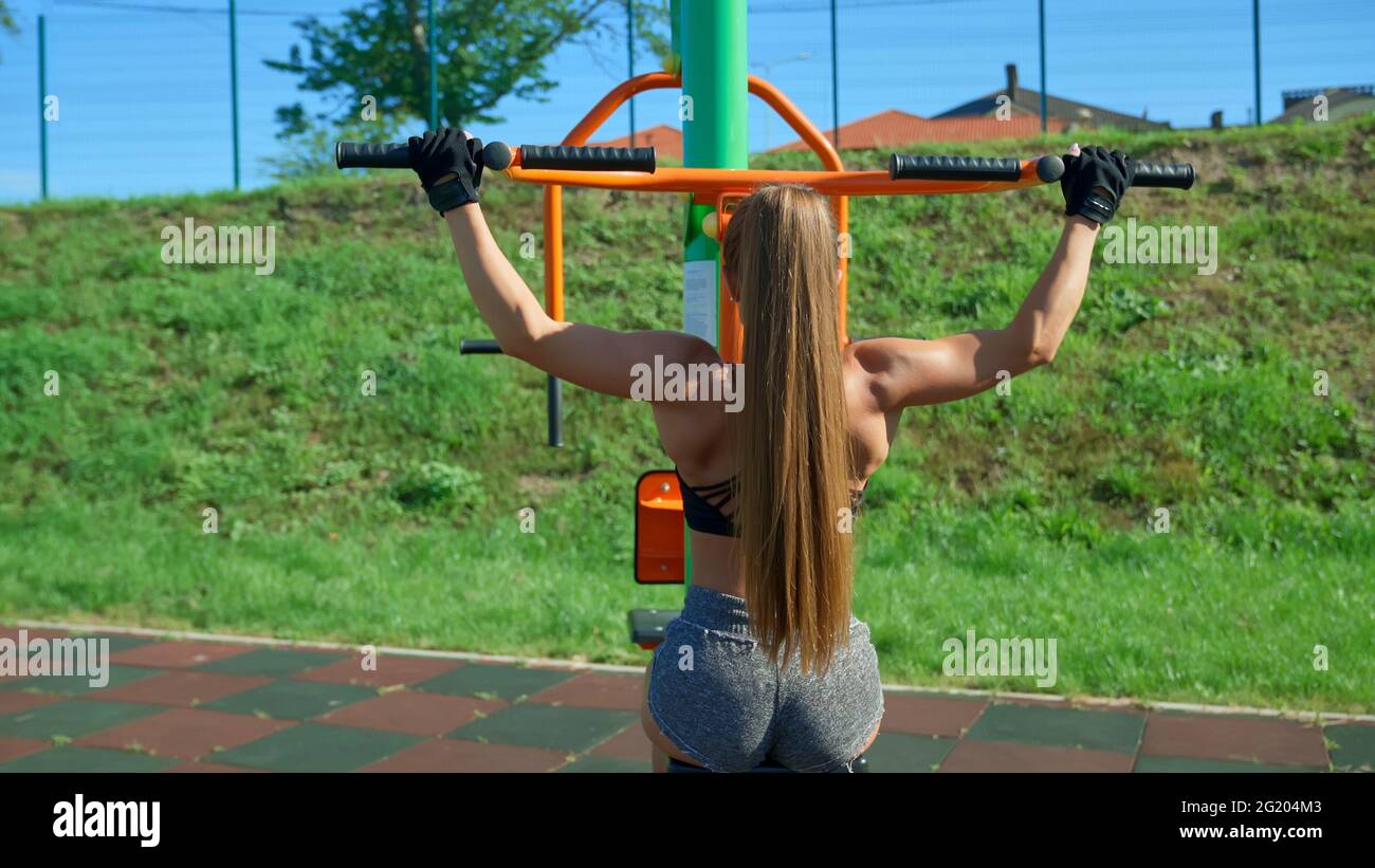 Young unrecognizable woman practicing wide grip lat pull downs using  simulator at sports ground outdoors. Back view of fit girl with perfect  muscular body and ponytail training back in sunny morning Stock