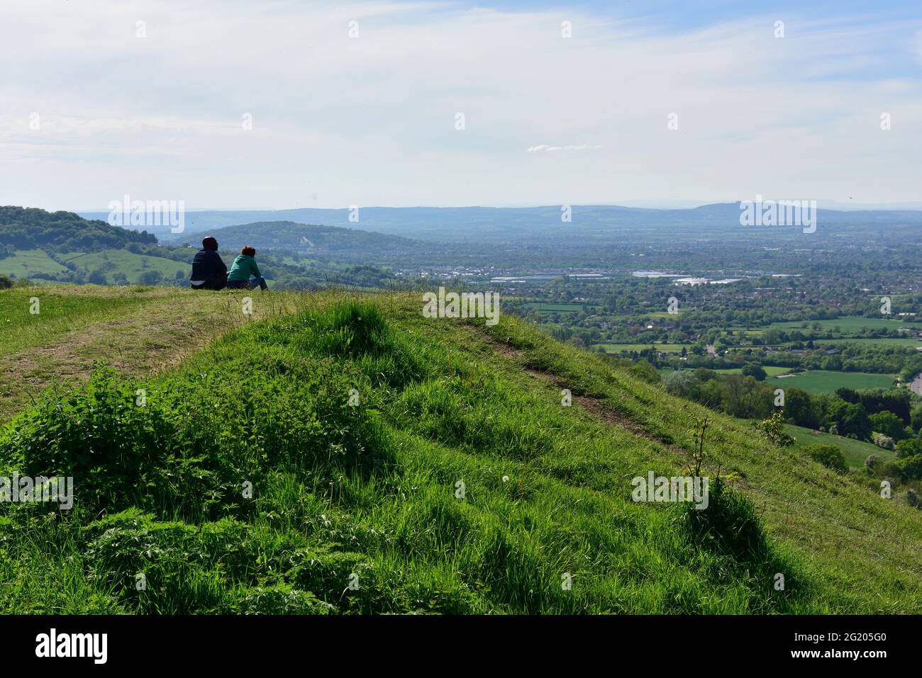 Two people sitting on grassy escarpment above Severn Vale looking across Gloucester toward Malvern Hills, Gloucestershire, UK Stock Photo