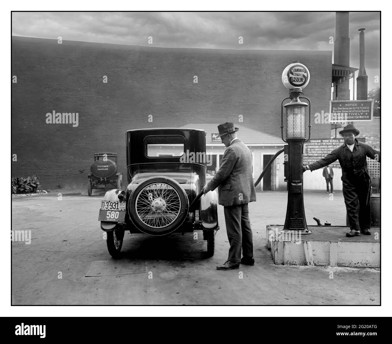 Archive 1900’s Gasoline Petrol Gas Station Man Fueling His Car At A Self-service Standard Motor gas station in Washington D.C. area. A sign tells customers the gasoline is filtered and contains no dirt or water. 1920. Washington USA Stock Photo