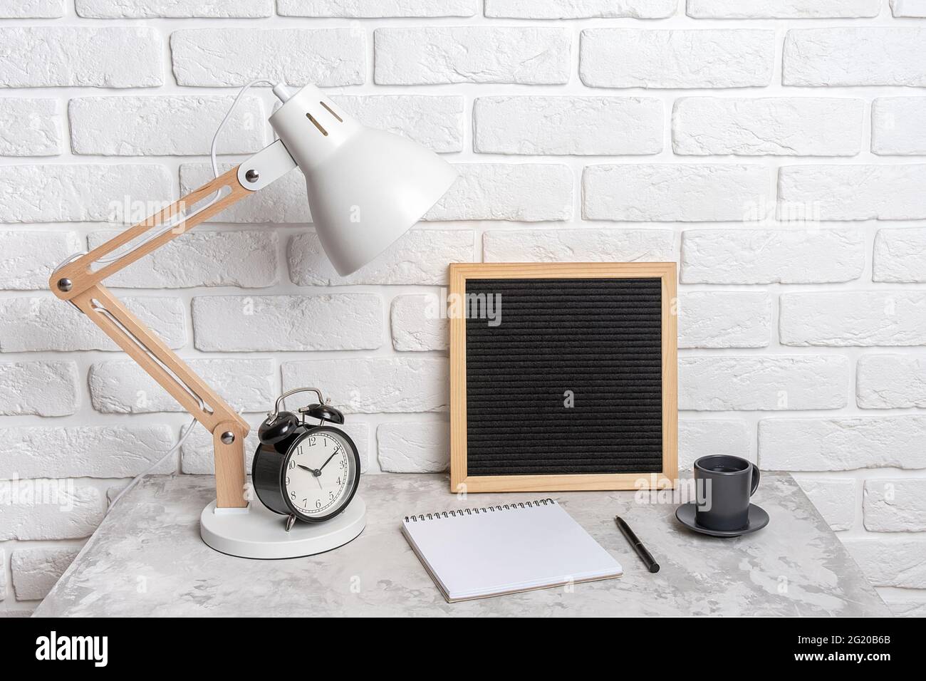 Table lamp, empty blank letter board, notebook and alarm clock on the table, against the background of a white brick wall. Workplace, front view, mock Stock Photo