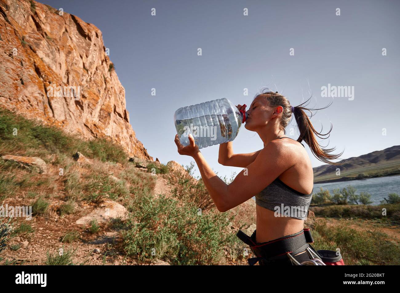 Young climber woman is drinking water from big huge bottle near the rocks and river at sunset. Tiredness and thirst concept. Stock Photo