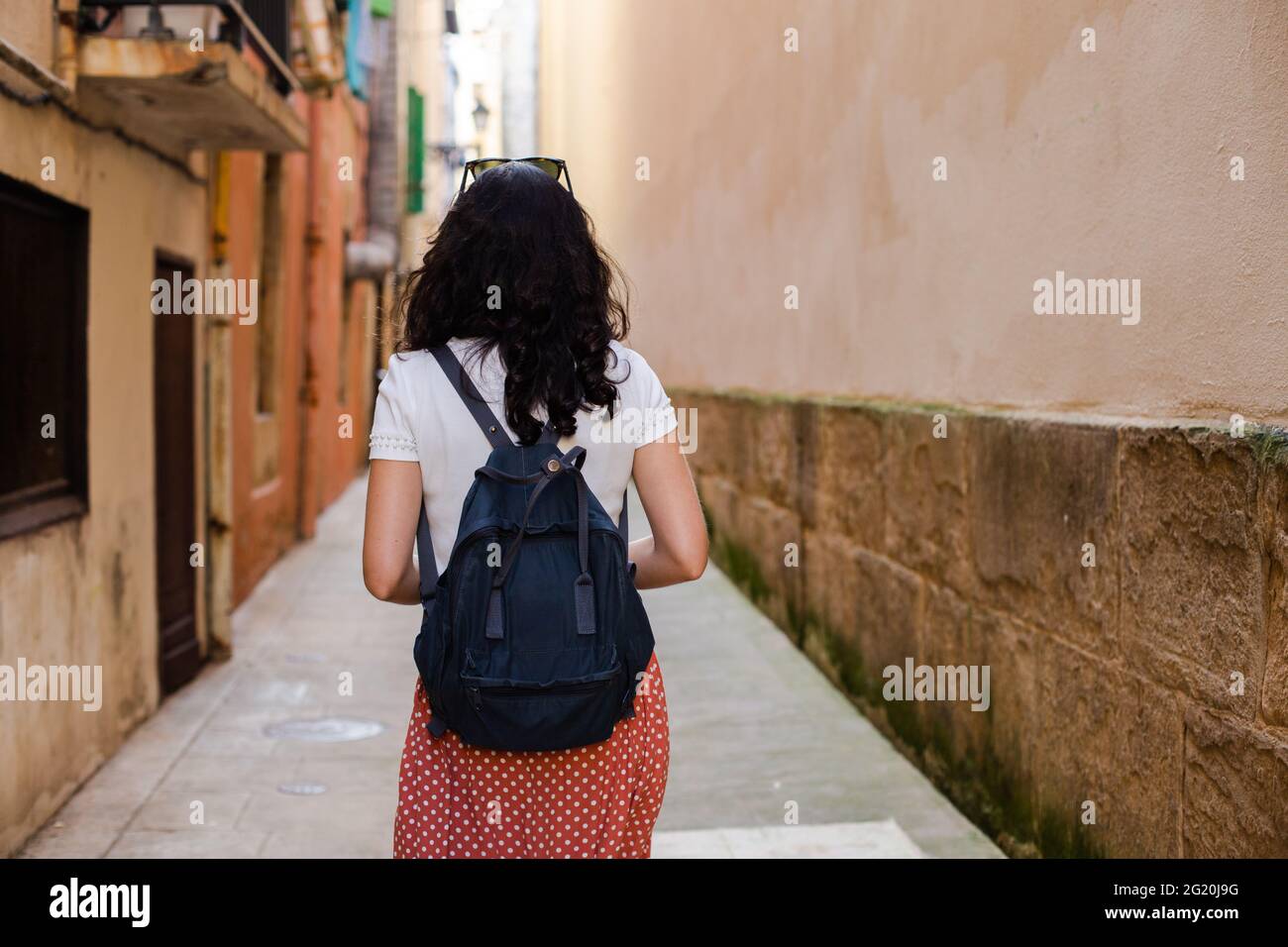 Rear view of a young woman with a backpack walking down a lonely city alley alone. Sense of danger Stock Photo
