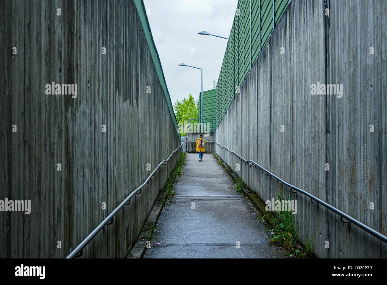 A woman in a yellow rain jacket walks from a station through a narrow footpath with high stone walls to the street. Stock Photo