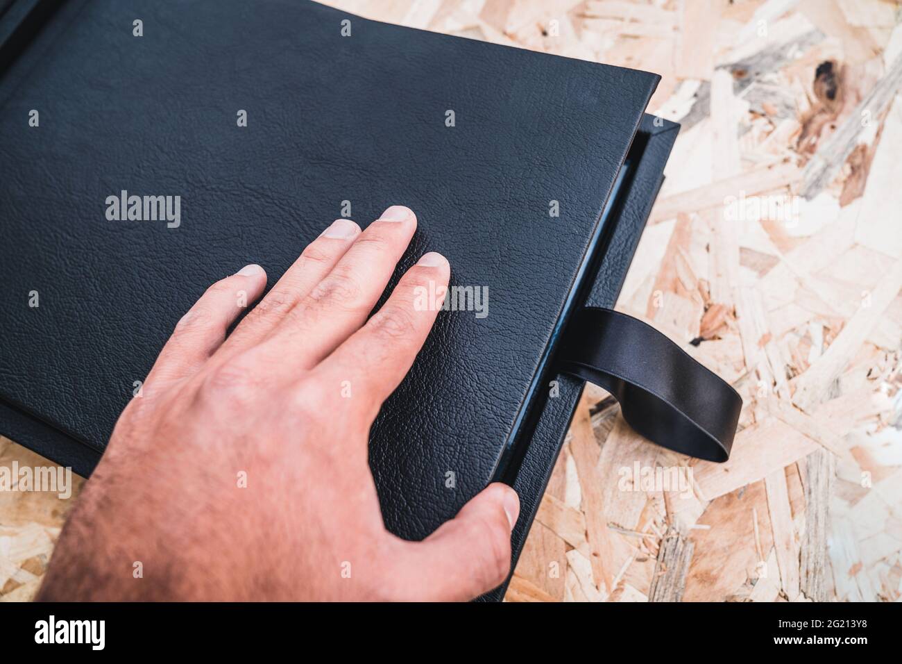 Anonymous person opening a black leather photo album by hand on a table with vintage wood background. Wedding or family photo book memories with a nic Stock Photo