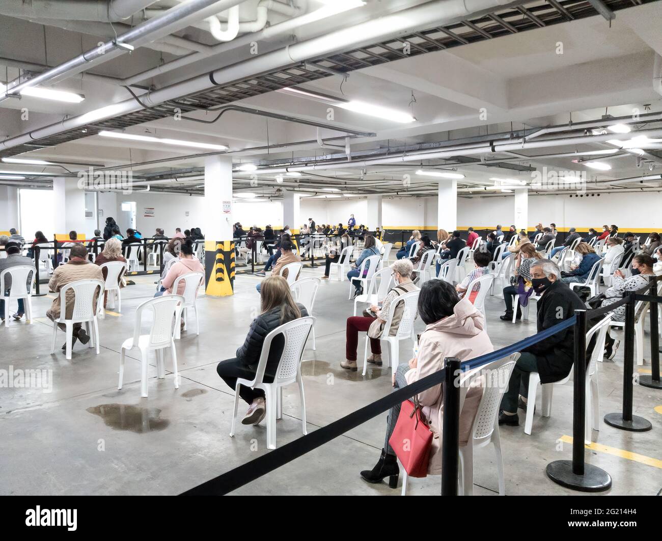Bogota, Colombia - June 03, 2021: Local Colombian people seen sitting on socially distanced plastic chairs and waiting in a public underground car park, to be vaccinated against Covid-19. They are all wearing protective face masks due to the pandemic situation. At a street level altitude of about 8,500 feet above mean sea level on the Andes Mountains, all people in the image are seen wearing warm clothing. Image taken on a mobile phone. Stock Photo