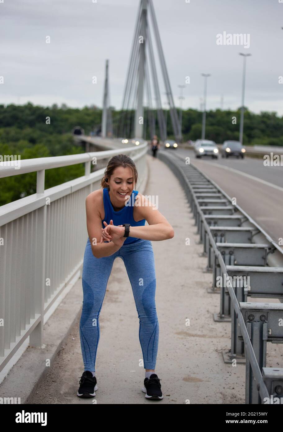 Fit girl bending setting smart watch on bridge. Young athlete jogging exercising. Stock Photo