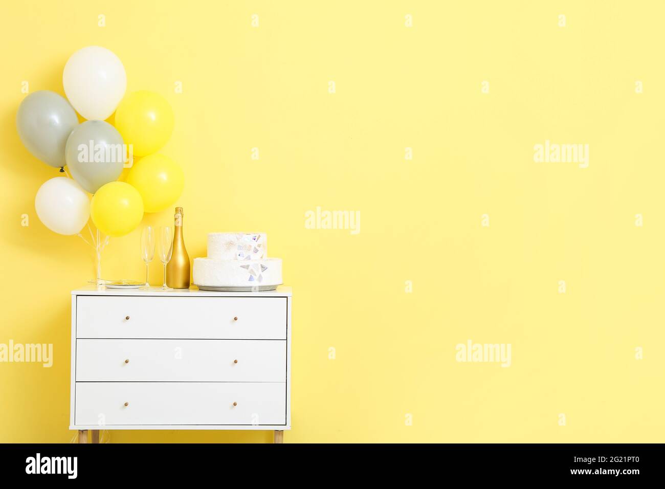 Birthday cake with champagne on chest of drawers and air balloons near color wall Stock Photo