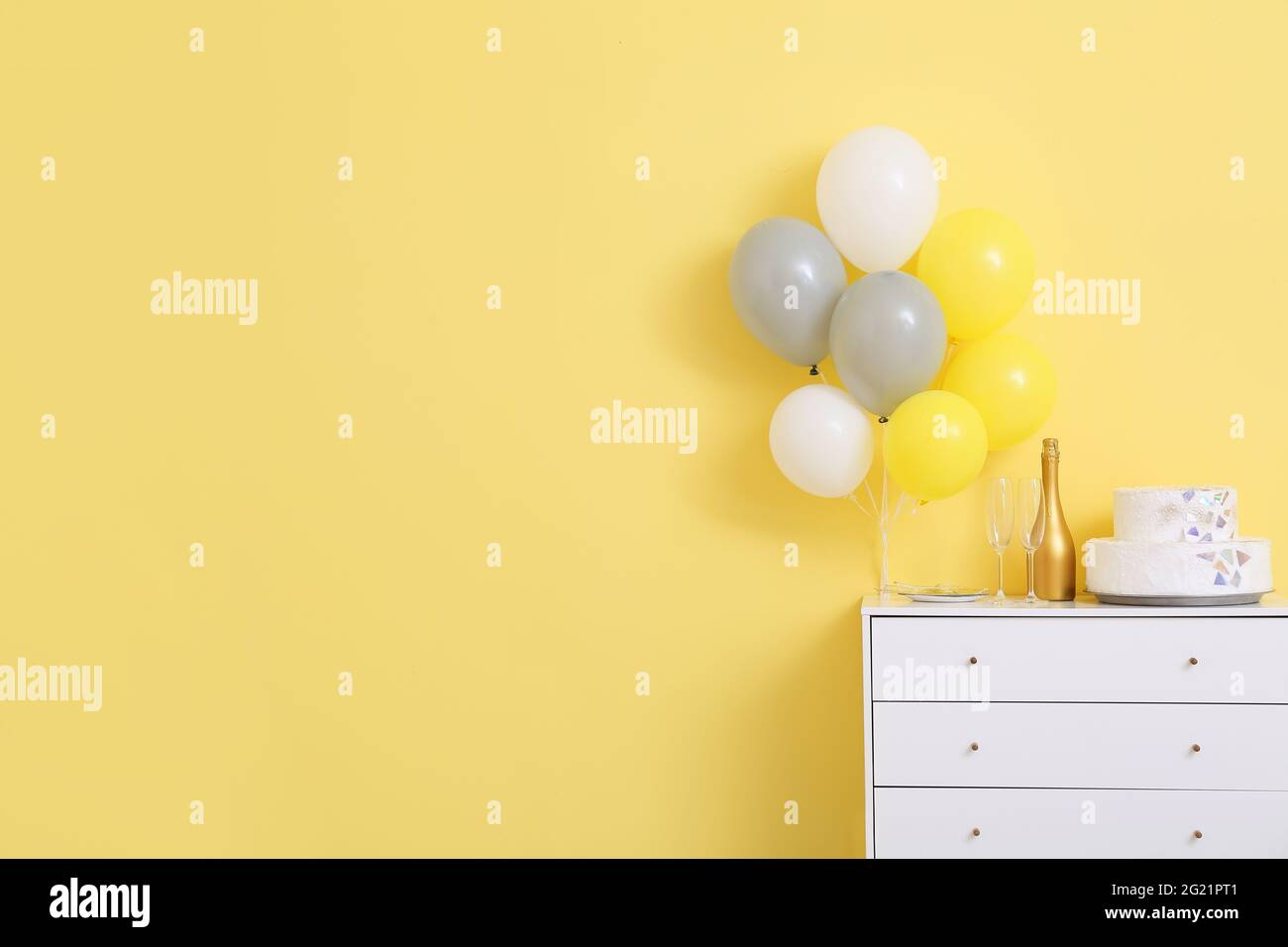 Birthday cake with champagne on chest of drawers and air balloons near color wall Stock Photo