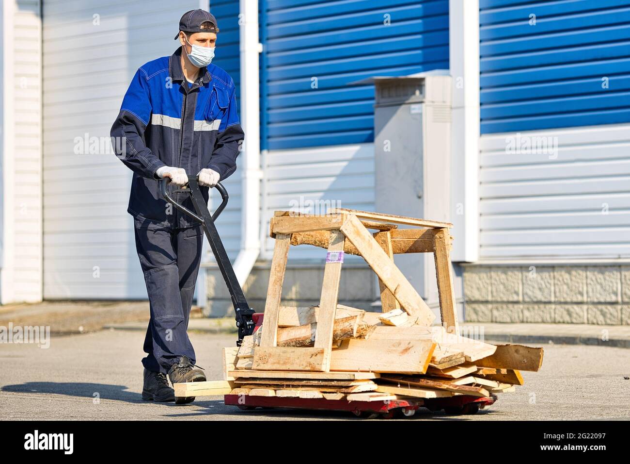 A man in work clothes and a medical mask carries wooden planks on a cart on the territory of an industrial base on a summer day. Work in accordance with sanitary standards Stock Photo