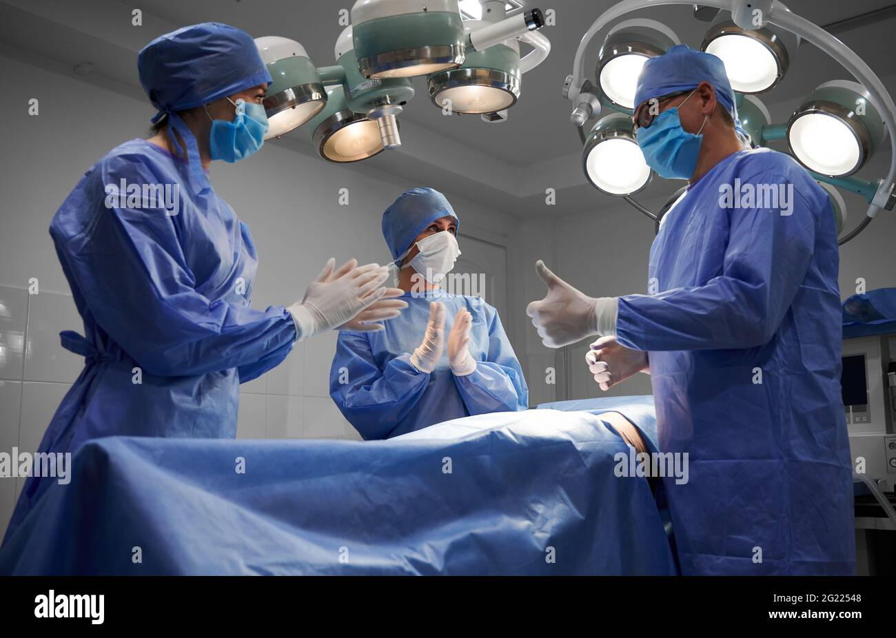 Doctors standing by patient after surgical operation. Surgeon doing thumbs up gesture while female assistants looking at man and applauding. Concept of medicine, plastic surgery, successful teamwork. Stock Photo