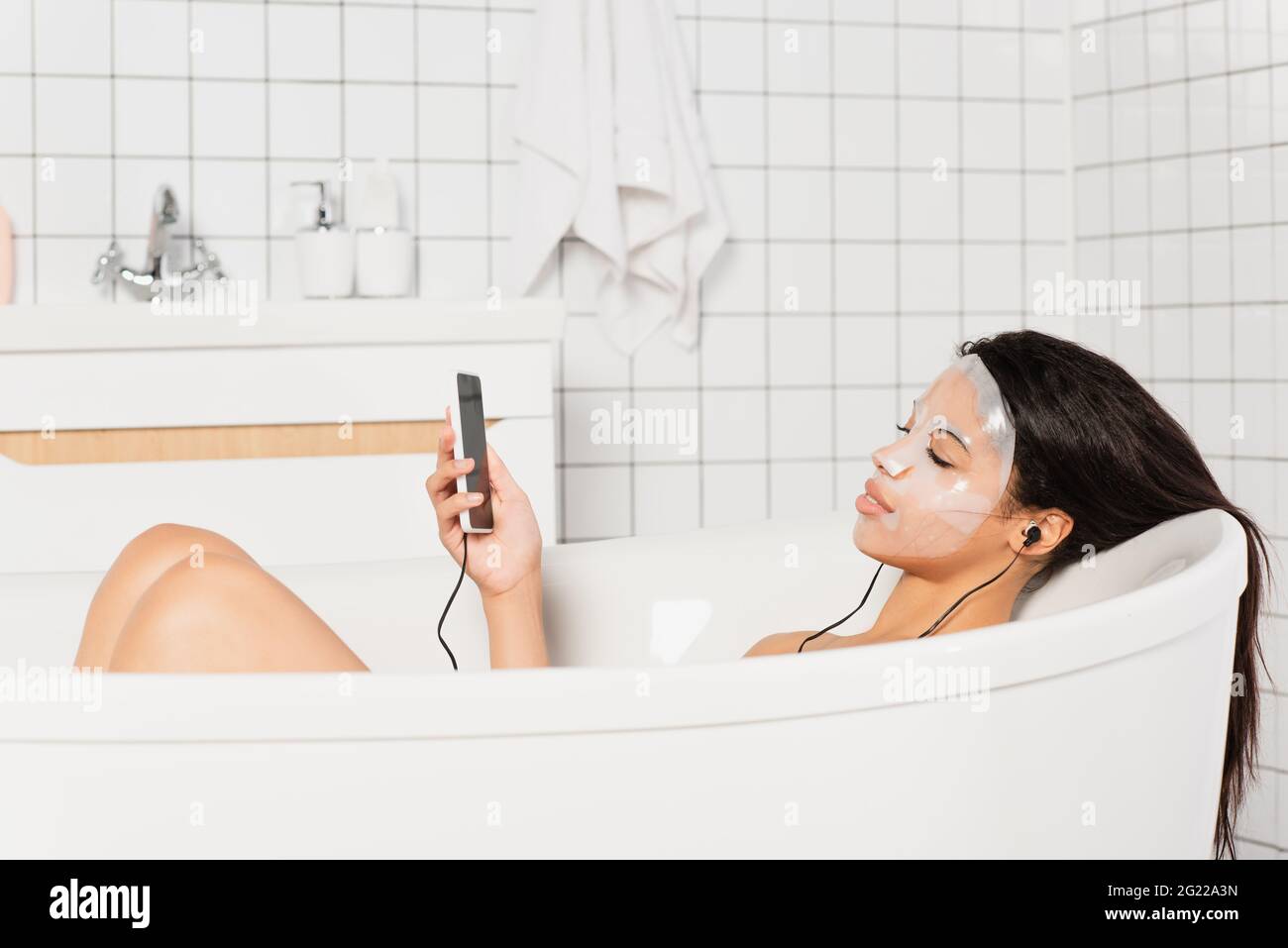 young woman with sheet mask on face lying with earphones and cellphone