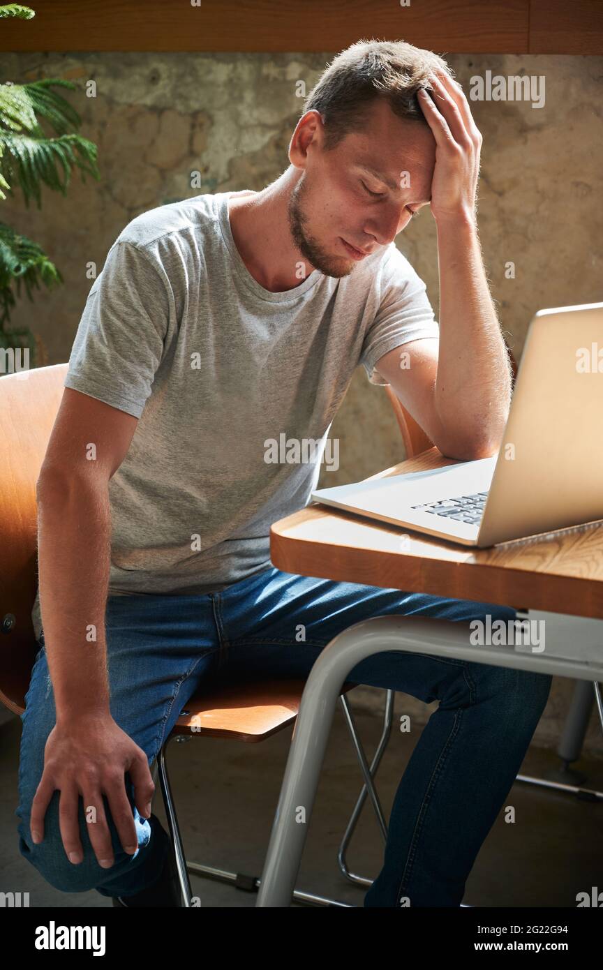 Young frustrated man in grey t-shirt sitting on laptop and thinking about work. Concept of process working in laptop at home or cafe. Stock Photo