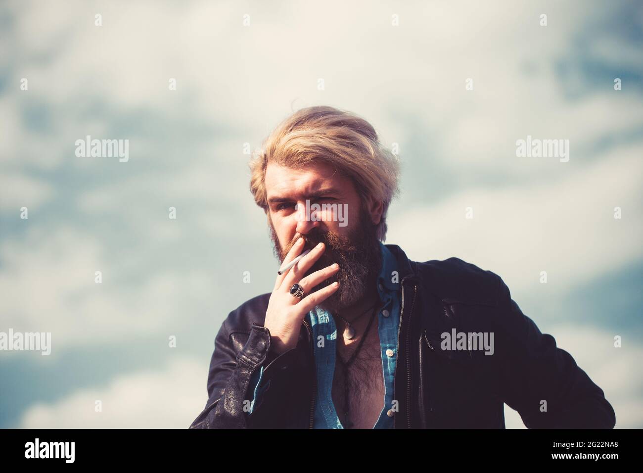 Trendy cool young man standing outside smoking cigarette. Against blue sky. Stock Photo