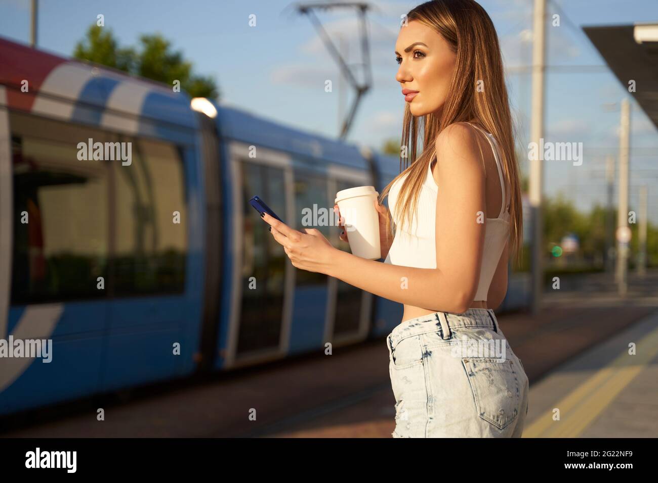 Young woman waiting for city train and talking on phone with coffee in hand. Passenger at streetcar stop with smartphone. Concept of public transport and communication. High quality photo  Stock Photo
