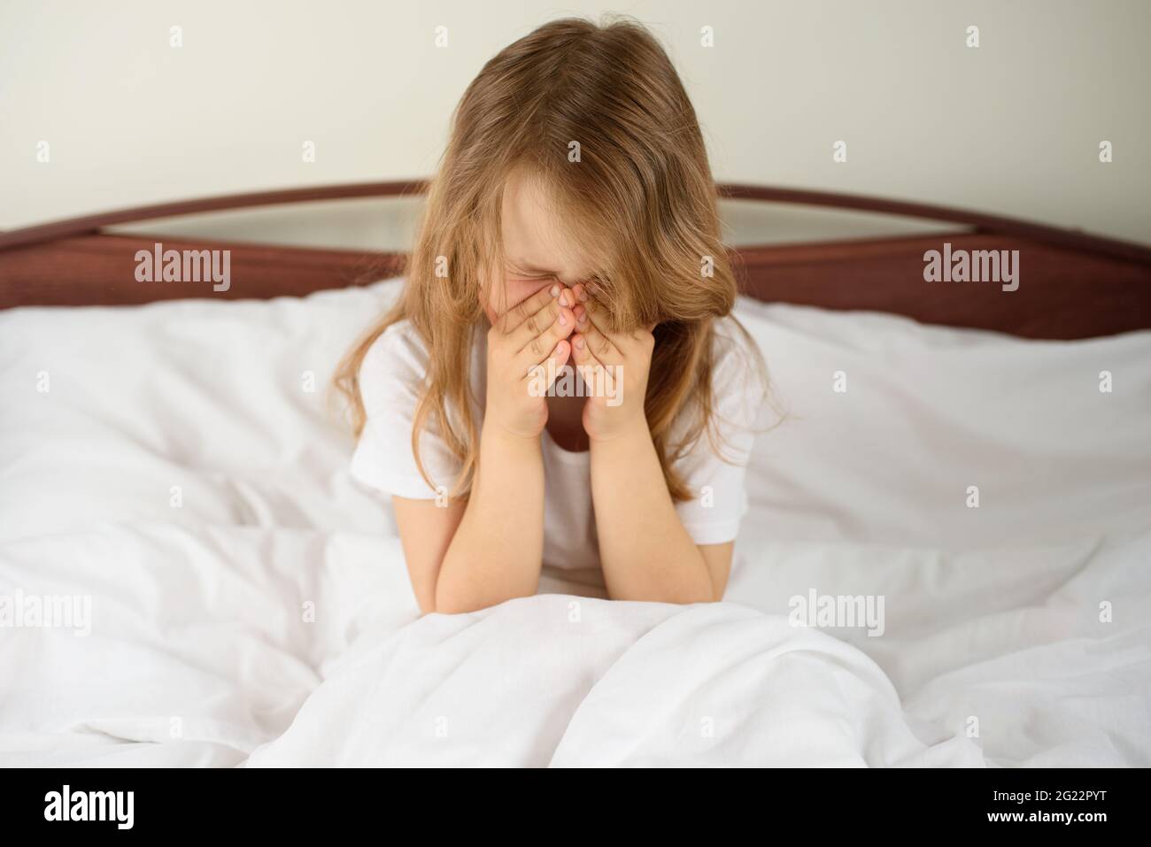 A little girl sits and sneezes on the bed among the blankets and pillows. House dust mite