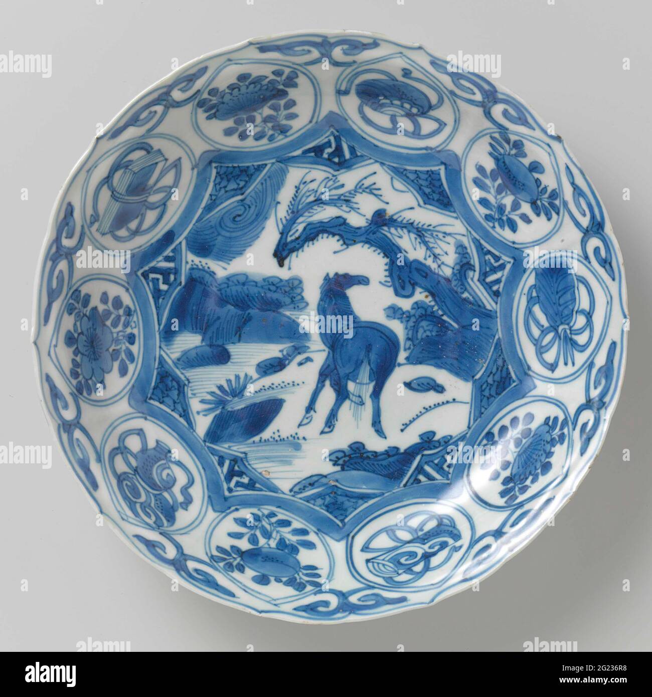 Dish. Scale of porcelain with a scalloped edge, painted in underglaze blue. On the flat a star-shaped cartouche with a horse in it, shortened from the rear, in a landscape with rocks and trees. Cartouche corners with napkin. The edge is divided into medallions with flower branches or lucky symbols (book, pearl, artemisia leaf, role, balebass jar); Between the medallions. The outer wall is divided into compartments with dots. Cracking porcelain in blue and white. Stock Photo